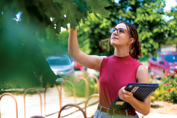 Ava Hartman, wearing glasses and a red shirt while holding a tablet, looks up to examine a tree's leaves.