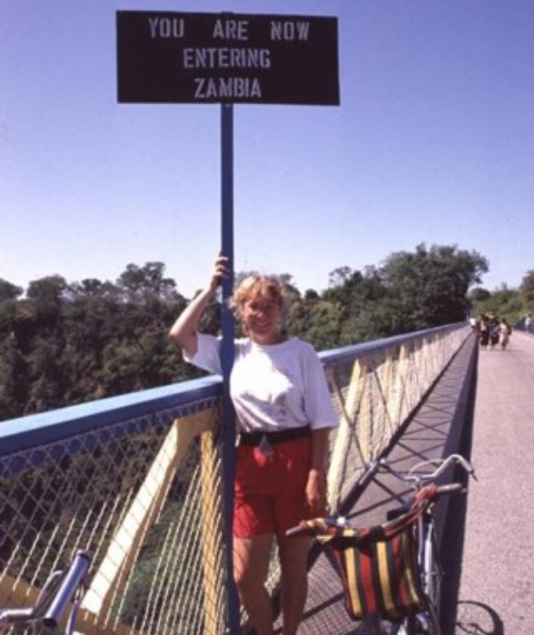 Susan Hume standing next to a sign that reads: You are now entering Zambia