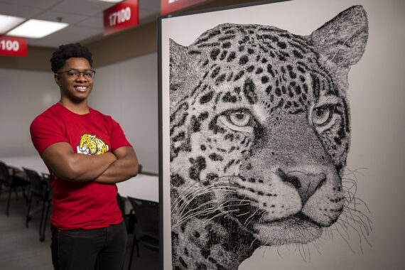 William Minion, wearing a red IUPUI Jaguars shirt, smiles while standing beside his black-and-white whiteboard drawing of a jaguar head.
