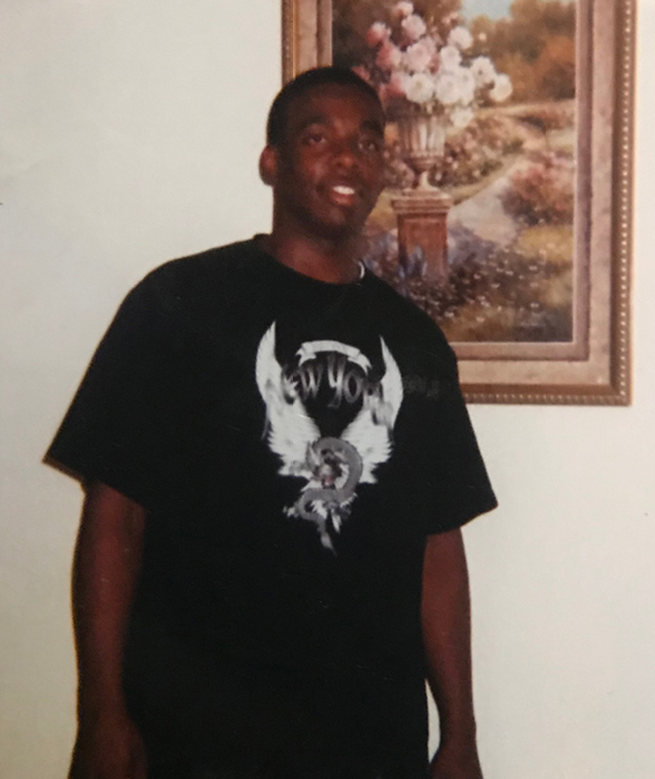 Russell Ledet smiles as he stands inside his childhood home. He’s wearing a black oversized shirt with a “New York” graphic on the front. Behind him hangs an artistic painting of a garden with a flower bouquet mounted in the center. 