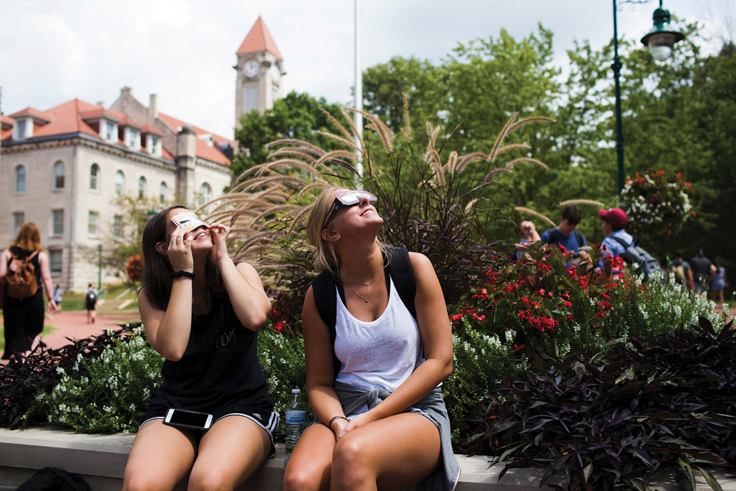 Two women in shorts and tank-tops—one wearing special glasses, the other holding a special viewing card to her face—smile as they look to the sky. They're sitting on the IU Bloomington campus in front of a flower bed full of plants, and the Student Building behind them in the distance.