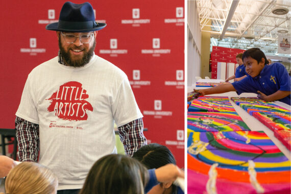 J.C. Barnett III, who has medium-brown skin and a dark-brown beard, wears clear-frame glasses and a black fedora, and a white T-shirt that says "Little Artists" on it. A young buy with medium-brown skin and wearing a blue polo shirt places a painted canvas on a table along with several other bright, colorful paintings.
