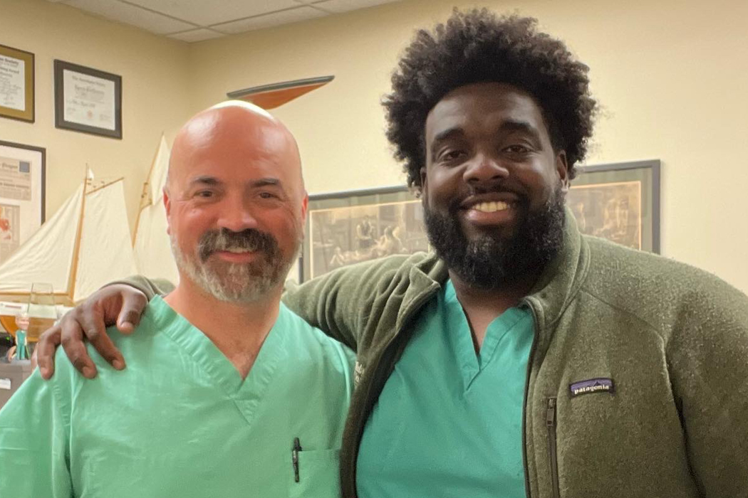 Two doctors stand beside each other inside a hospital room. On the left is Patrick Greiffenstein, Caucasian with a white and black beard and mustache. The second is Russell Ledet, a Black American with a black afro and beard. His arm lays around Patrick Greiffenstein's shoulder in a friendly embrace. They are both wearing seafoam green scrubs. 
