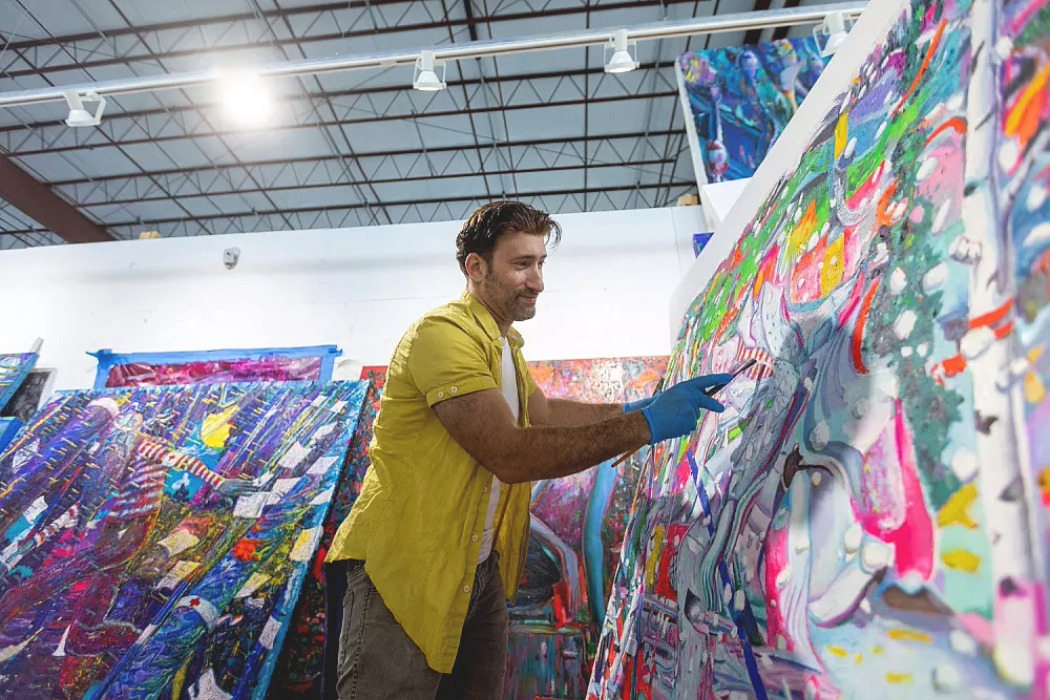In a brightly lit studio with large canvases stacked against the walls, a man with tan skin and brown hair wearing a short-sleeve, yellow button-up shirt and blue gloves holds a paintbrush up to a canvas full of undulating, overlapping colorful shapes.